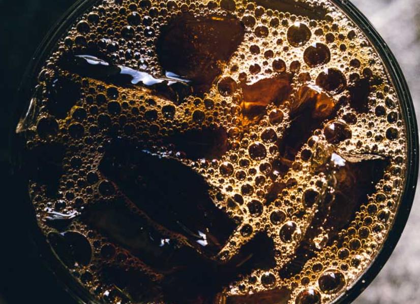 The image shows a close-up of a glass filled with iced coffee. The dark brown liquid is topped with numerous ice cubes, and bubbles have formed on the surface, creating a rich texture. 