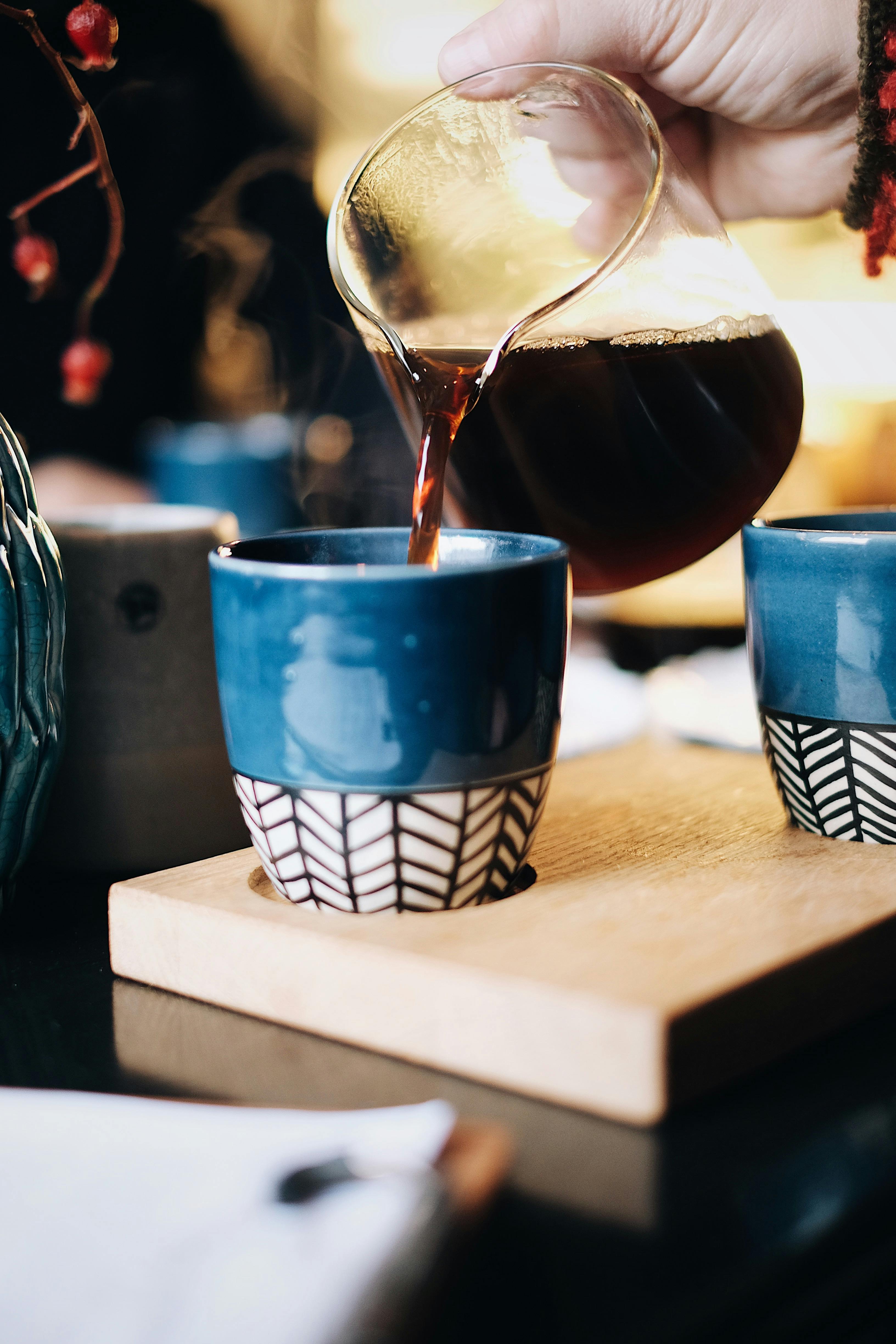 This image captures a close-up of a hand pouring freshly brewed coffee from a glass carafe into a blue ceramic cup with a black and white geometric pattern at the bottom. 