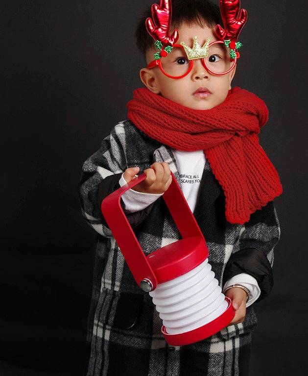 Child holding a red foldable LED night light, dressed in festive winter attire. Portable USB rechargeable lamp with touch dimmable brightness.