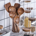 Teak wood kitchen set from Thailand featuring spoons, ladles, and turners in a glass measuring cup. Durable and eco-friendly tableware for cooking.