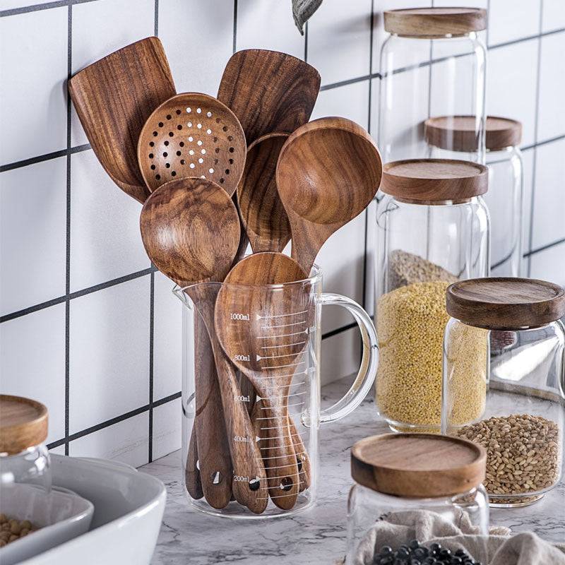 Teak wood kitchen set from Thailand featuring spoons, ladles, and turners in a glass measuring cup. Durable and eco-friendly tableware for cooking.