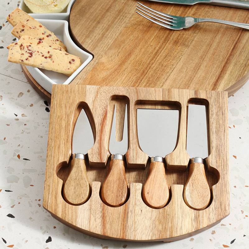 Wooden cheese board with four cheese knives on a speckled surface.