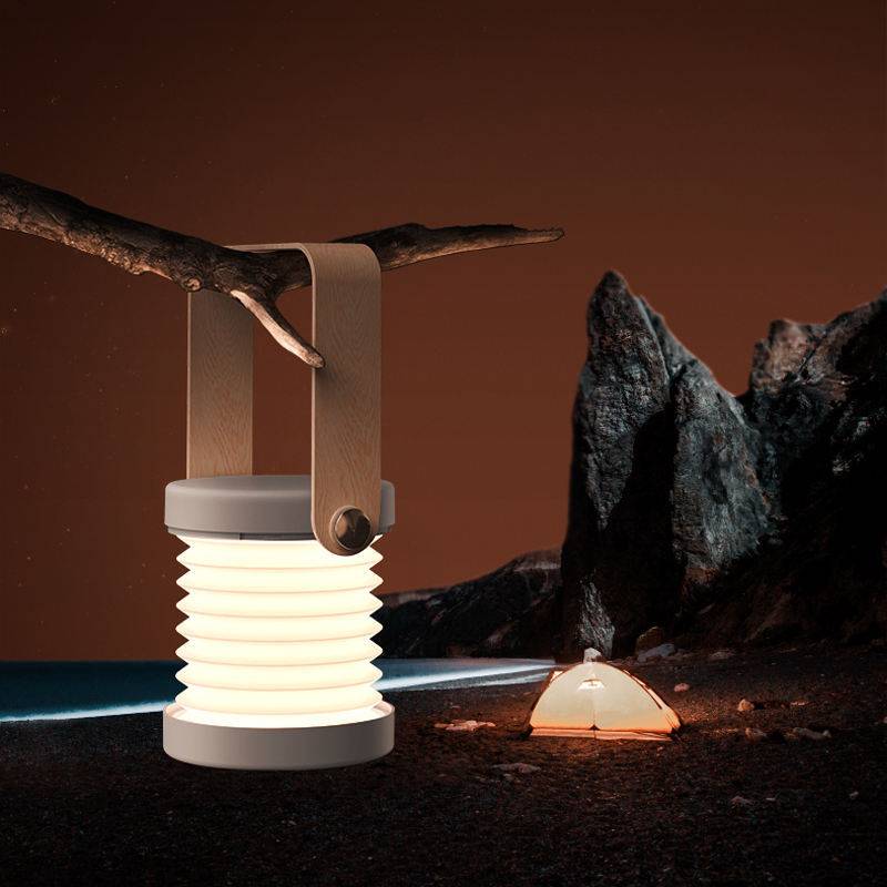 Foldable LED night light hanging from a tree branch, illuminating an outdoor camping scene. USB rechargeable lantern with touch dimmable control.