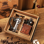 Coffee-making set with French press and grinder in a box on a wooden surface with coffee beans.