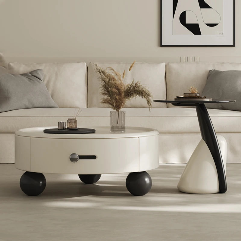 A modern white coffee table with black accents and gold legs, round in shape, with a book and a vase of dried flowers on top, placed in a minimalist living room setting.