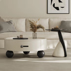 A modern white coffee table with black accents and gold legs, round in shape, with a book and a vase of dried flowers on top, placed in a minimalist living room setting.
