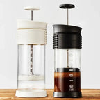 Two coffee presses, one white and one black with a textured handle, on a wooden surface.