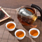 Stainless steel tea infuser steeping in a glass teapot with three cups of tea. Ideal for brewing loose leaf tea with rich flavor.