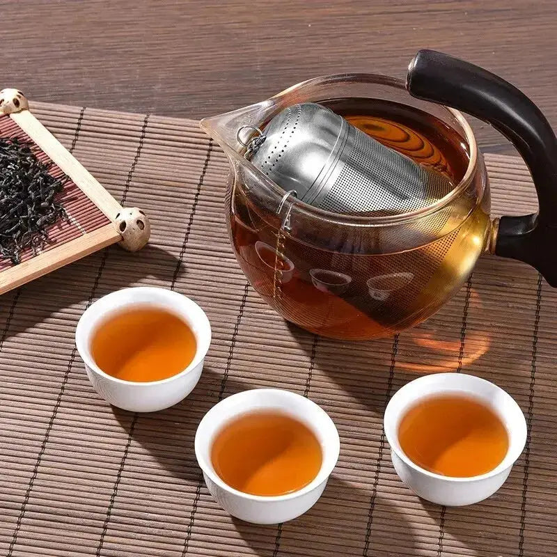 Stainless steel tea infuser steeping in a glass teapot with three cups of tea. Ideal for brewing loose leaf tea with rich flavor.