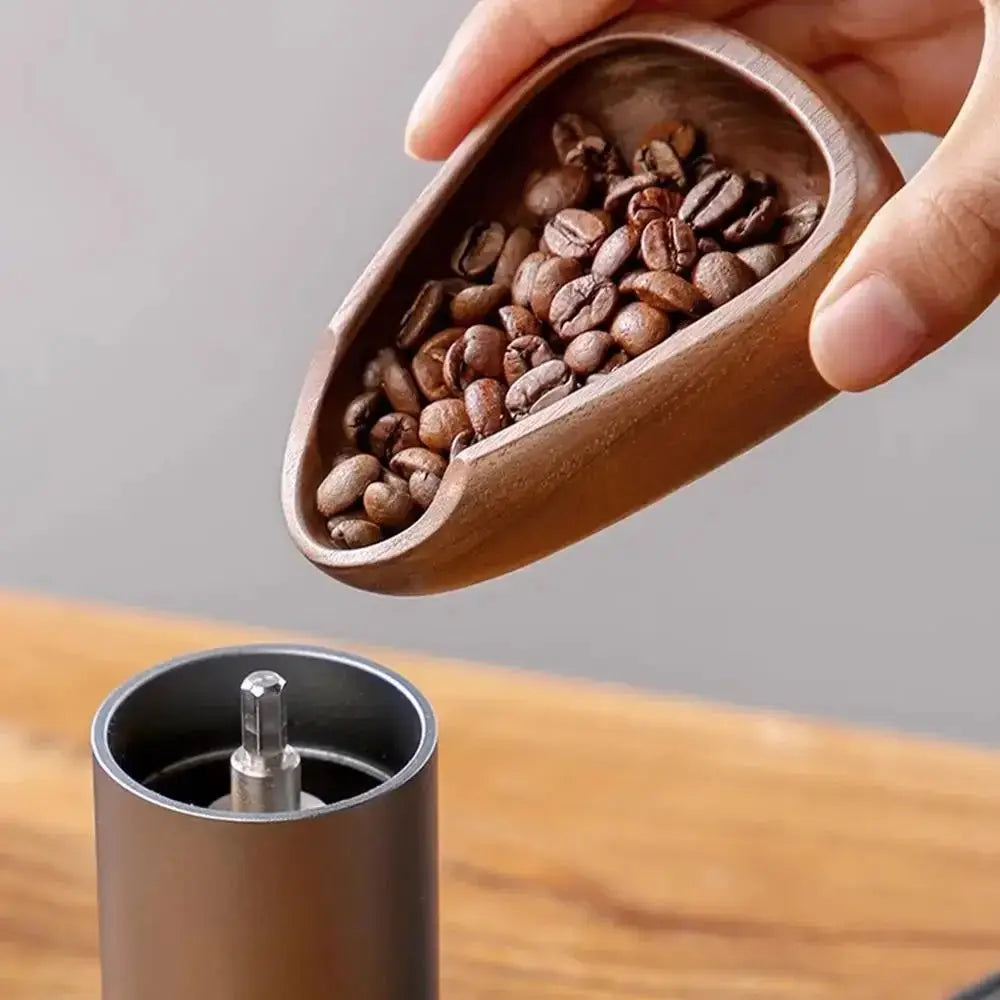 Hand holding a wooden scoop filled with coffee beans above a coffee grinder.