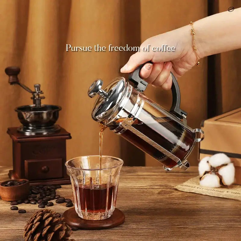 Hand pouring coffee from a French press into a glass on a wooden table with a coffee grinder and beans in the background.