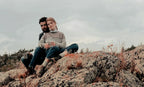 Couple sitting on rocky terrain, one holding a portable espresso maker, with a scenic outdoor backdrop and cloudy sky.