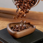Coffee beans being poured from a transparent container into a wooden bowl on a wooden surface.