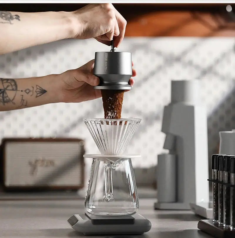 Freshly ground coffee being poured into a pour-over dripper with glass carafe on counter