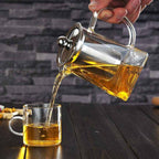 Glass infuser teapot with stainless steel lid and filter, pouring freshly brewed tea into a matching glass cup. Ideal for effortless tea brewing.